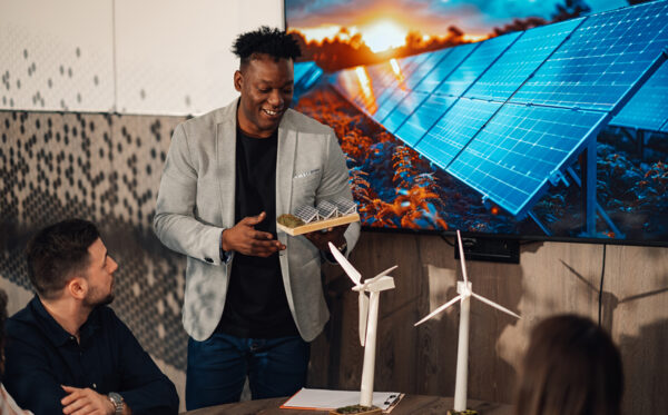 Diverse businessman with solar panel model having seminar at boa Portrait of multicultural businessman standing at meeting room with solar panels miniature in hands and having presentation on a briefing. Smiling ecologist having seminar about green technology.
